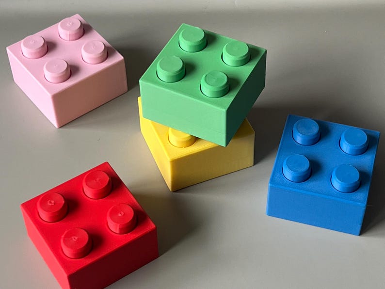 May include: A collection of colorful building block toys in pink, green, yellow, blue, and red. Each block has four raised circular studs on top. The blocks are arranged on a light gray surface.