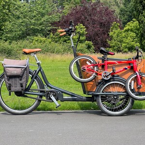 May include: A black cargo bike with a brown wooden box and a red folding bike attached. The bike has a brown leather seat and a gray pannier bag. The background features green trees and grass.