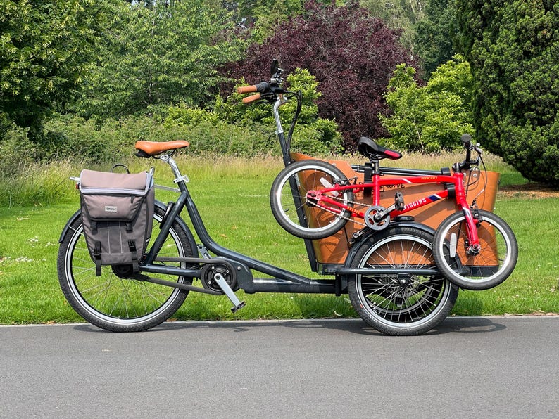 May include: A black cargo bike with a brown wooden box and a red folding bike attached. The bike has a brown leather seat and a gray pannier bag. The background features green trees and grass.