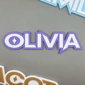 May include: A layered, three-dimensional sign with the name "OLIVIA" in white letters, outlined in a light purple color. The letter "O" has a diamond shape in the center. Other name signs are visible.