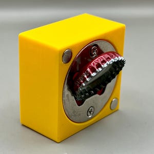 Op de afbeelding: Een felgele flesopener met een zilveren cirkelvormige opening. Een rode flesdop is gedeeltelijk ingebracht. De opener heeft een kubusvorm met afgeronde zilveren klinknagels aan de zijkanten.
