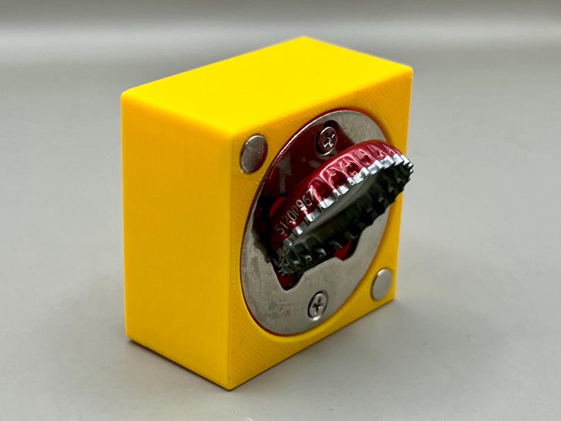 Op de afbeelding: Een felgele flesopener met een zilveren cirkelvormige opening. Een rode flesdop is gedeeltelijk ingebracht. De opener heeft een kubusvorm met afgeronde zilveren klinknagels aan de zijkanten.