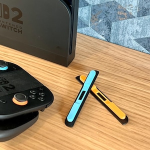 May include: A black Nintendo Switch console and controller are shown, alongside two coloured Joy-Con rail covers. One cover is light blue, the other is orange. The covers are designed to enhance the look of the gaming console.