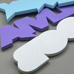 May include: Close-up of colorful foam shapes in light blue, purple, and white. The shapes are geometric and appear to be part of a larger design or puzzle. The foam has a textured surface, and the background is a solid gray.