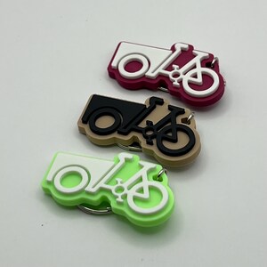 May include: Three keychains shaped like a bicycle with a cargo box. The keychains are in three different colors: pink, brown, and green. The bicycle and cargo box are white on each keychain.