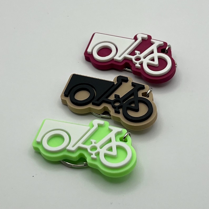 May include: Three keychains shaped like a bicycle with a cargo box. The keychains are in three different colors: pink, brown, and green. The bicycle and cargo box are white on each keychain.