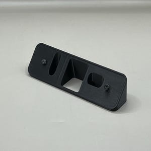 May include: A black rectangular plastic object with two screw holes and a central opening. The object has a raised section on one side and is likely a mounting bracket or holder.