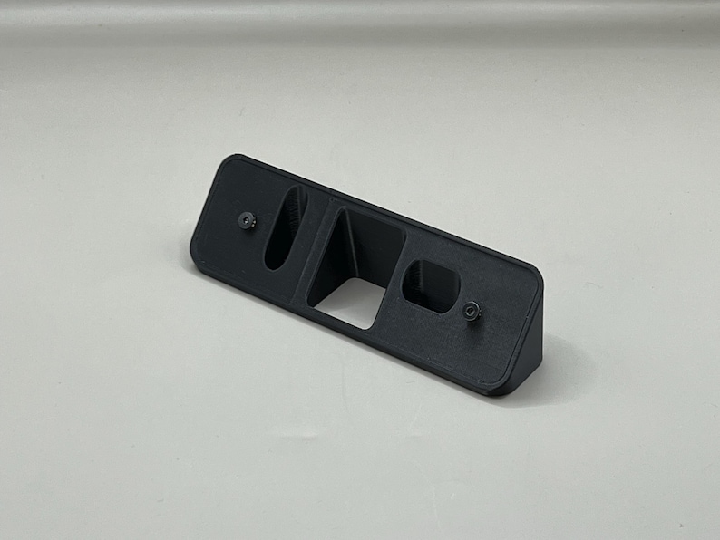 May include: A black rectangular plastic object with two screw holes and a central opening. The object has a raised section on one side and is likely a mounting bracket or holder.