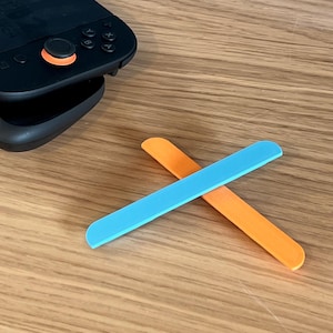 May include: A pair of colorful, rectangular game controller accessories. One is bright blue, the other is orange. They are positioned in an X shape on a wooden surface, near a black game controller.