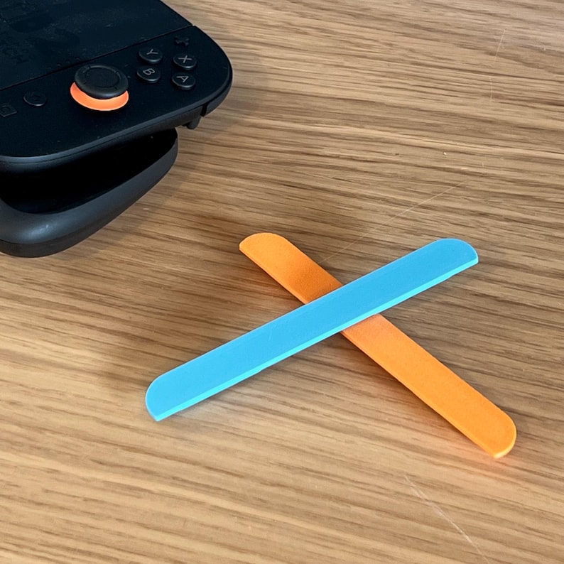 May include: A pair of colorful, rectangular game controller accessories. One is bright blue, the other is orange. They are positioned in an X shape on a wooden surface, near a black game controller.