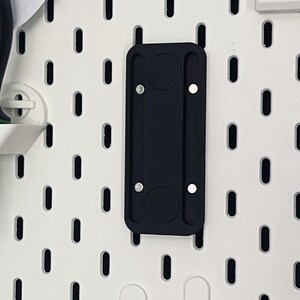 Peut inclure: Un accessoire noir pour panneau perfor&eacute; avec deux renfoncements circulaires et deux points de montage argent&eacute;s. L'accessoire rectangulaire a des bords arrondis et est mont&eacute; sur un panneau perfor&eacute; blanc avec des trous ovales.