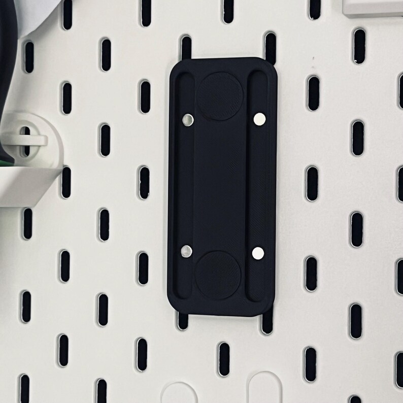 Peut inclure: Un accessoire noir pour panneau perfor&eacute; avec deux renfoncements circulaires et deux points de montage argent&eacute;s. L'accessoire rectangulaire a des bords arrondis et est mont&eacute; sur un panneau perfor&eacute; blanc avec des trous ovales.