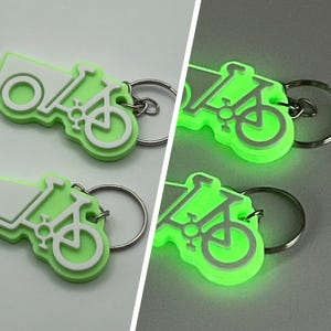 May include: Two green glow-in-the-dark bike keychain charms with white bike silhouettes. The charms are attached to silver key rings.