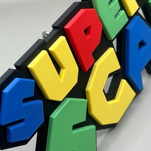 Puede incluir: Decoraci&oacute;n mural colorida con la palabra "SUPER" en letras 3D. Las letras son de colores brillantes: amarillo, rojo, azul y verde, sobre un fondo negro. Las letras en relieve crean un efecto dimensional. Dise&ntilde;ado para colgar en la pared.