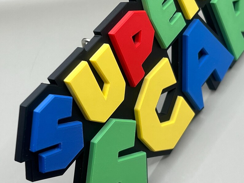 Puede incluir: Decoraci&oacute;n mural colorida con la palabra "SUPER" en letras 3D. Las letras son de colores brillantes: amarillo, rojo, azul y verde, sobre un fondo negro. Las letras en relieve crean un efecto dimensional. Dise&ntilde;ado para colgar en la pared.