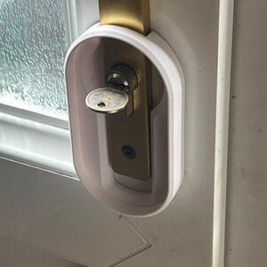 May include: A white door lock cover installed on a white door. The cover surrounds a brass-colored door lock with a key inserted. The door has a glass window panel to the left.