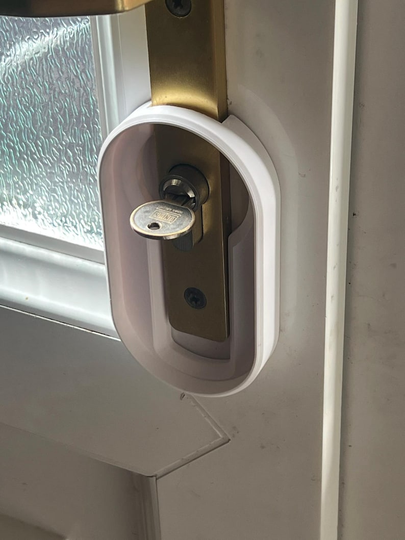 May include: A white door lock cover installed on a white door. The cover surrounds a brass-colored door lock with a key inserted. The door has a glass window panel to the left.
