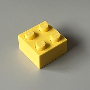May include: A bright yellow building block with four raised studs on top. The block is square-shaped and sits on a gray surface. The studs are the same yellow color as the block.
