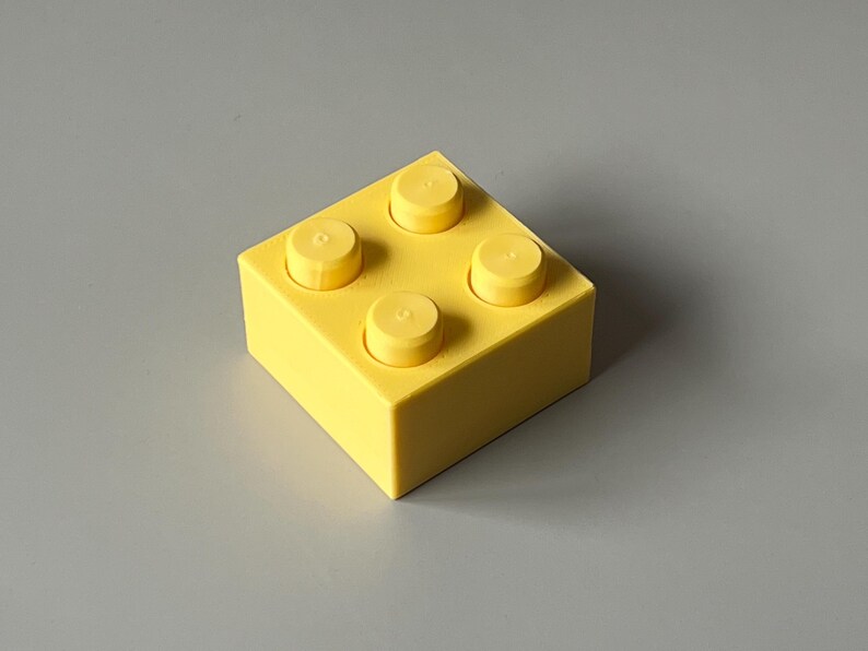 May include: A bright yellow building block with four raised studs on top. The block is square-shaped and sits on a gray surface. The studs are the same yellow color as the block.