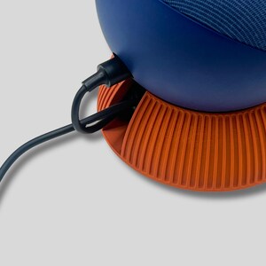 Puede incluir: Primer plano de un altavoz inteligente con una parte superior azul marino y una base naranja. El altavoz tiene un cable de alimentaci&oacute;n negro enchufado en la parte posterior. La base tiene crestas verticales.