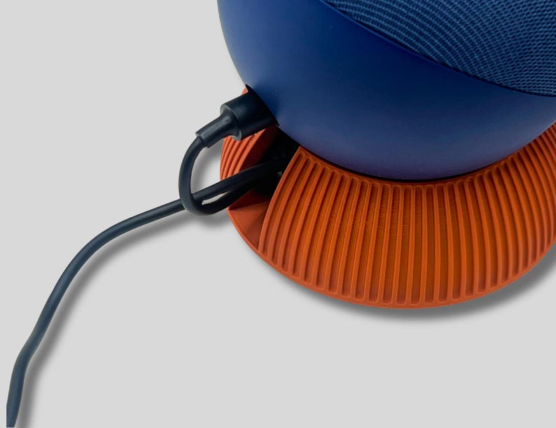 Puede incluir: Primer plano de un altavoz inteligente con una parte superior azul marino y una base naranja. El altavoz tiene un cable de alimentaci&oacute;n negro enchufado en la parte posterior. La base tiene crestas verticales.