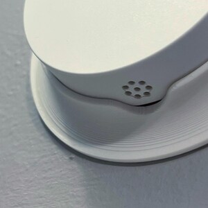 May include: Close-up of a white smoke detector mounted on a light gray wall. The smoke detector has a circular base with concentric ridges and a rounded top with small ventilation holes.