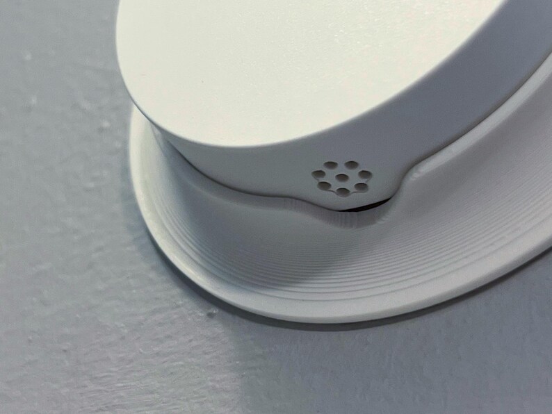 May include: Close-up of a white smoke detector mounted on a light gray wall. The smoke detector has a circular base with concentric ridges and a rounded top with small ventilation holes.