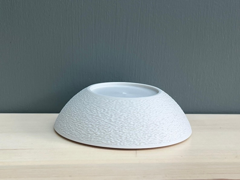 May include: A white, shallow, circular bowl with a textured exterior surface featuring a fine, rippled pattern. The bowl has a smooth, flat interior base and a wide, rounded rim, presenting a clean, monochromatic appearance throughout the object.