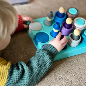 May include: A child plays with a wooden toy sorting game. The game features a blue tray with various sized and colored wooden circles and figures. The child is placing a blue circle into the tray.