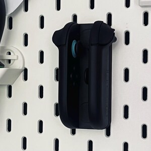 Peut inclure: Un support de manette noir mont&eacute; sur un panneau perfor&eacute; blanc. Le support est con&ccedil;u pour maintenir solidement une manette de jeu, avec un joystick bleu visible. Le panneau perfor&eacute; comporte des trous ovales espac&eacute;s.