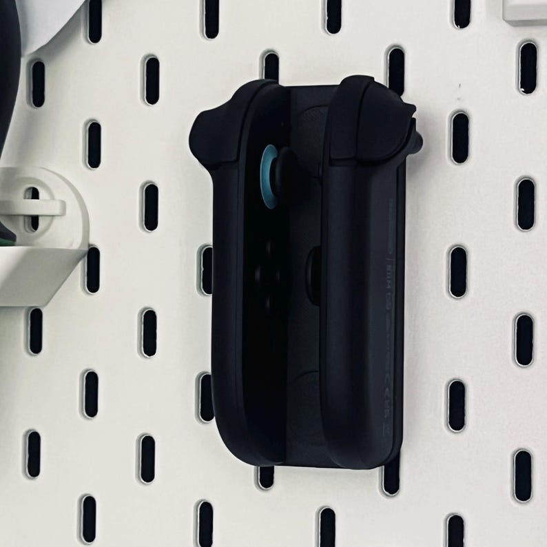 Peut inclure: Un support de manette noir mont&eacute; sur un panneau perfor&eacute; blanc. Le support est con&ccedil;u pour maintenir solidement une manette de jeu, avec un joystick bleu visible. Le panneau perfor&eacute; comporte des trous ovales espac&eacute;s.