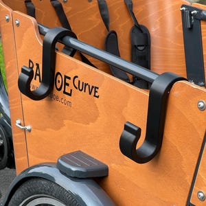May include: Close-up of a wooden cargo bike with black metal accents. The bike features a wooden cargo box with the text "BA OE Curve" printed on the side. Black hooks and a gray metal bar are visible.