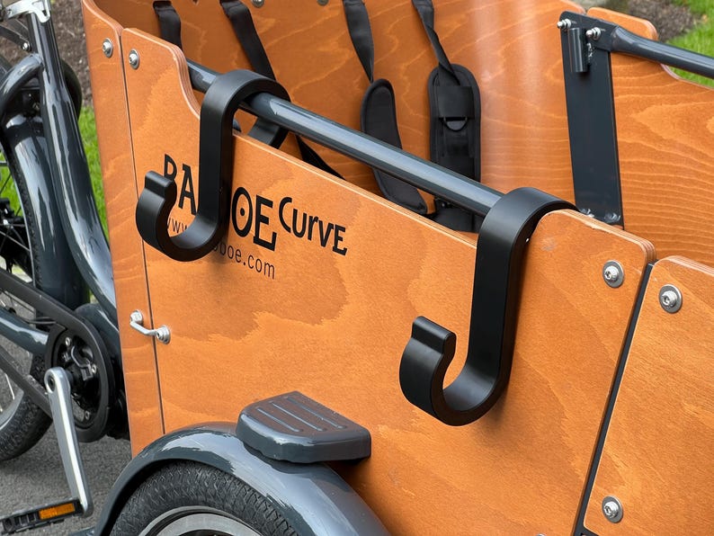 May include: Close-up of a wooden cargo bike with black metal accents. The bike features a wooden cargo box with the text "BA OE Curve" printed on the side. Black hooks and a gray metal bar are visible.