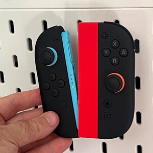 Puede incluir: Mandos Joy-Con de Nintendo Switch negros y rojos. El mando izquierdo es negro con un acento azul, y el mando derecho es negro con un acento rojo. Los mandos tienen botones y joysticks.