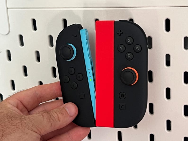 Puede incluir: Mandos Joy-Con de Nintendo Switch negros y rojos. El mando izquierdo es negro con un acento azul, y el mando derecho es negro con un acento rojo. Los mandos tienen botones y joysticks.