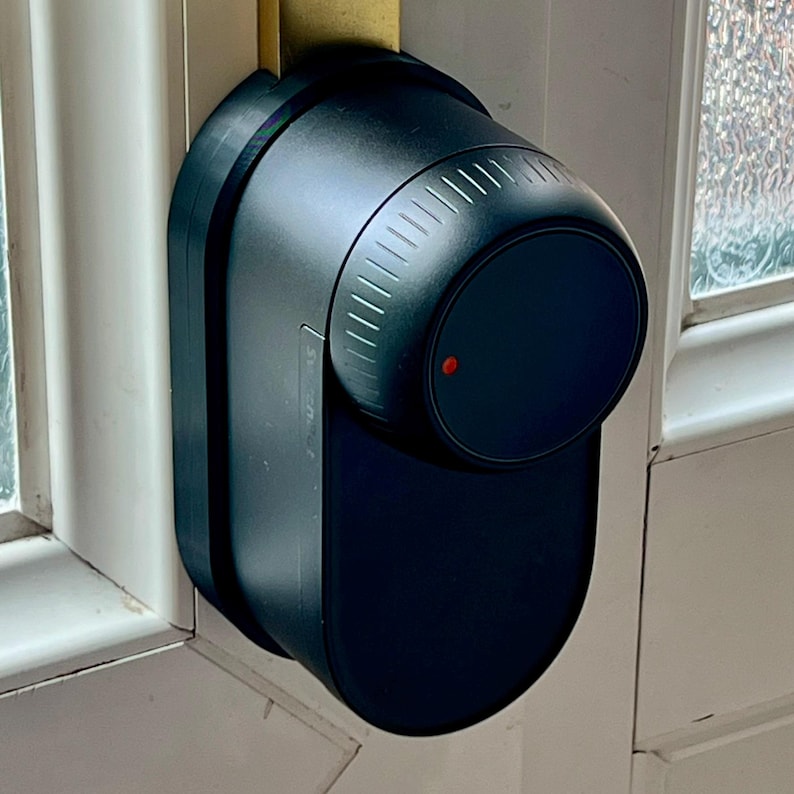 May include: A black smart lock installed on a white door. The lock has a cylindrical dial with vertical grooves and a small red indicator light. The lock's design is modern and sleek, with a focus on functionality and security.