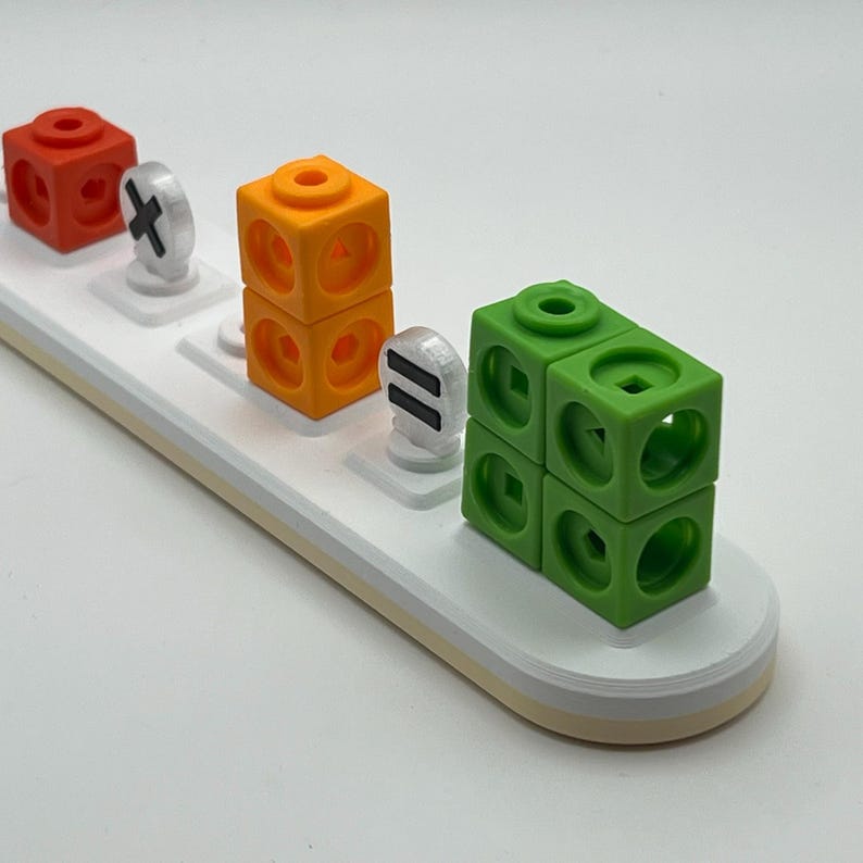 May include: A white plastic base with three slots for colorful plastic blocks. The blocks are stacked and feature different shapes. The first block is red, the second is orange, and the third is green. The slots have white plastic pieces with symbols: a plus sign, an equals sign, and a minus sign.