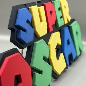 Puede incluir: Un letrero 3D multicolor que dice "SUPER OSCAR". Las letras son azules, amarillas, rojas y verdes, con un contorno negro. Probablemente sea un art&iacute;culo decorativo o un juguete.