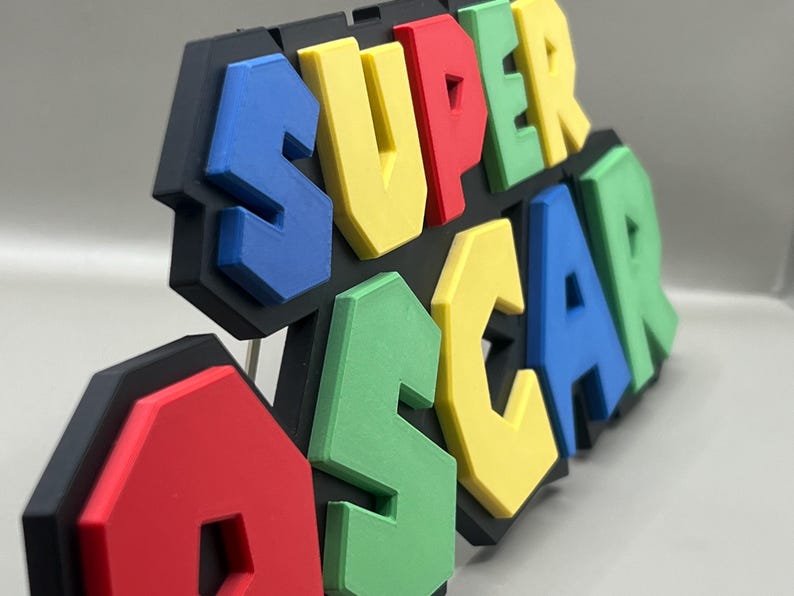 Puede incluir: Un letrero 3D multicolor que dice "SUPER OSCAR". Las letras son azules, amarillas, rojas y verdes, con un contorno negro. Probablemente sea un art&iacute;culo decorativo o un juguete.