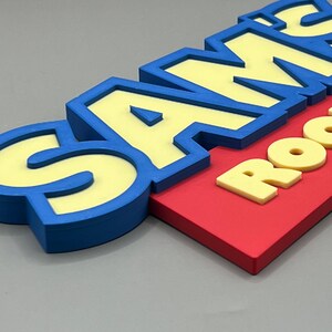 May include: A colorful sign that reads "SAM'S ROOM." The letters are yellow with blue outlines, set against a red background. The sign is designed with a 3D effect, creating a playful and eye-catching appearance.