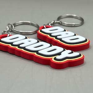 May include: Two keychains with the words "DADDY" and "DAD" in white, outlined in black and yellow, and set against a red background. Each keychain has a silver keyring attached.