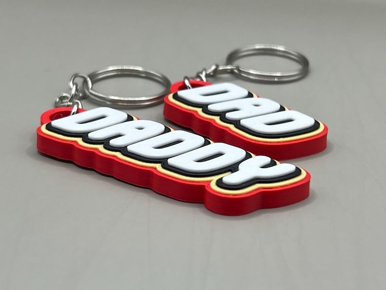 May include: Two keychains with the words "DADDY" and "DAD" in white, outlined in black and yellow, and set against a red background. Each keychain has a silver keyring attached.
