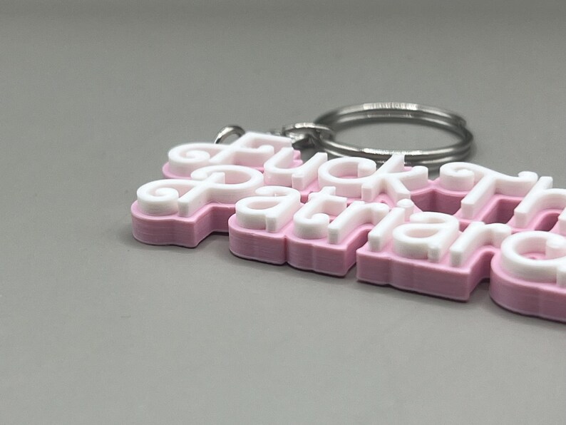 May include: A pink and white keychain with the words "Fuck Patriarchy" in a layered design. The keychain has a silver metal ring for attaching keys. The letters are white with a pink outline.