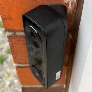 May include: A black rectangular security camera mounted on a red brick wall. The camera has a lens and a speaker. The camera is mounted on a white surface.