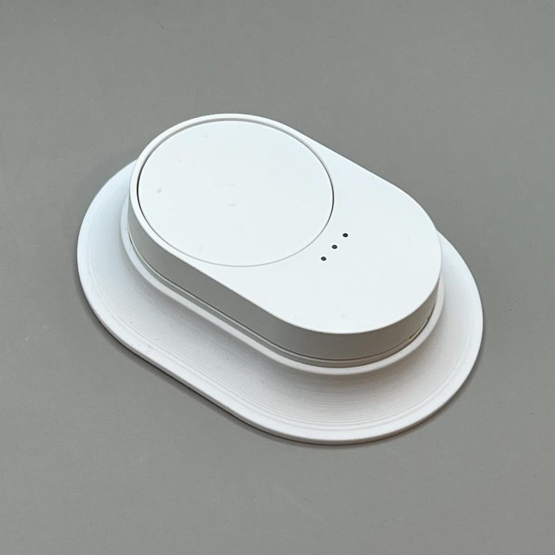 Pu&ograve; includere: Un pad di ricarica wireless bianco, di forma ovale, con una parte superiore circolare e tre piccoli punti indicatori. Poggia su una base bianca abbinata. Il design &egrave; pulito e minimalista. Il dispositivo &egrave; progettato per caricare dispositivi elettronici.