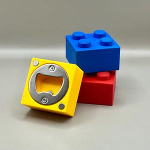 Op de afbeelding: Een gele flesopener in de vorm van een bouwsteen, met een zilveren opener, is gestapeld met blauwe en rode bouwstenen. De opener heeft een vierkante vorm met een metalen opener in het midden.