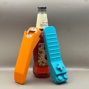 May include: An orange bottle opener and a blue bottle opener shaped like a video game controller are displayed in front of a bottle of red liquid. The bottle has a white label with text and a black cap.