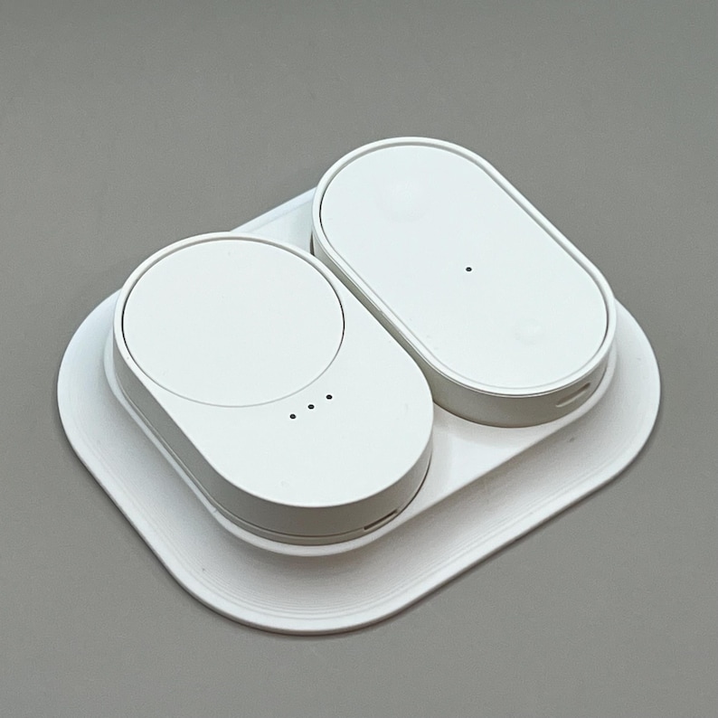 Pu&ograve; includere: Una stazione di ricarica wireless bianca con due basi di ricarica ovali e una base quadrata. Un pad ha la parte superiore circolare, l'altro &egrave; un ovale pieno. La stazione &egrave; progettata per caricare pi&ugrave; dispositivi contemporaneamente.