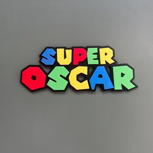 Puede incluir: Decoraci&oacute;n mural colorida con la inscripci&oacute;n "SUPER OSCAR" en una fuente l&uacute;dica y en bloques. Las letras son rojas, azules, amarillas y verdes con contornos negros, que recuerdan a un popular videojuego. La obra est&aacute; montada sobre un fondo gris.