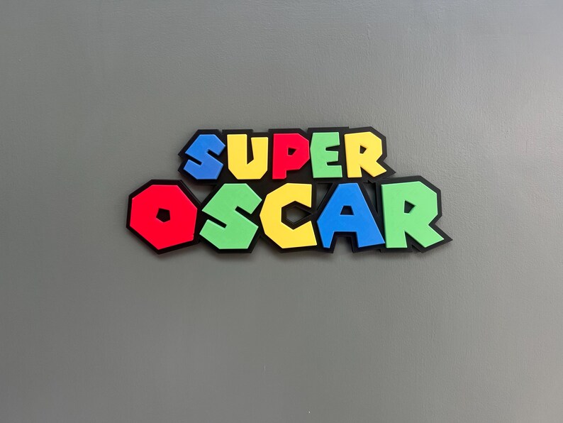 Puede incluir: Decoraci&oacute;n mural colorida con la inscripci&oacute;n "SUPER OSCAR" en una fuente l&uacute;dica y en bloques. Las letras son rojas, azules, amarillas y verdes con contornos negros, que recuerdan a un popular videojuego. La obra est&aacute; montada sobre un fondo gris.